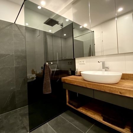 Dunkle Glasdusche hinter Waschbecken mit hölzernem Unterschrank und Spiegel