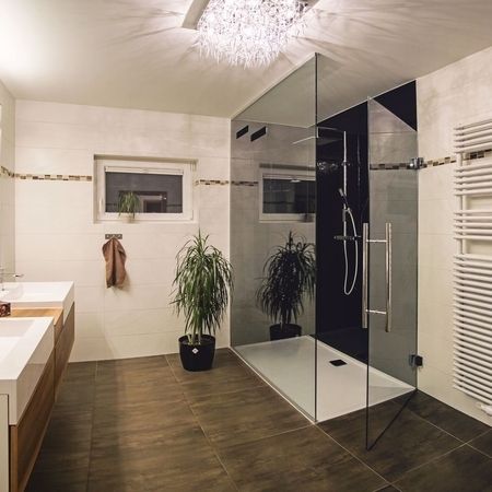 Bad mit braunem Boden, weißen Wandfliesen, einer Topfpflanze, Waschbecken links und einer Glasdusche rechts