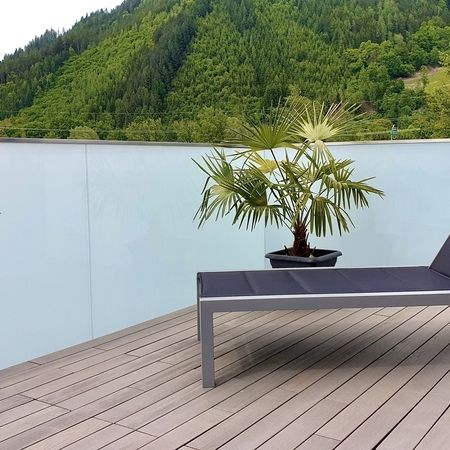 Milchglas-Terrassenverglasung mit Holzboden, Liege, Topfpalme und Naturhintergrund