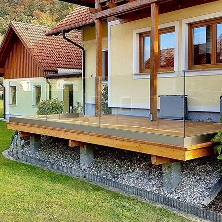 Glasgeländer auf erhöhter hölzerner Terrasse eines gelb-braunen Hauses, umgeben von einer Wiese und Bäumen und einem Haus im Hintergrund